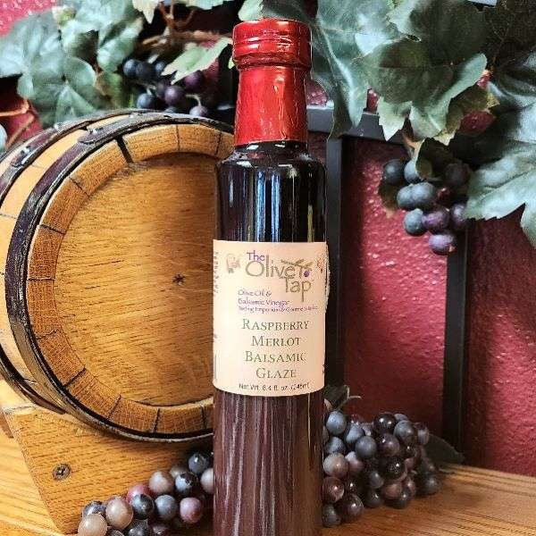 A bottle of Raspberry Merlot Balsamic Glaze from The Olive Tap in front of a decorative wooden barrel, purple grapes and their leaves, and a red wall in the background.