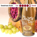 A bottle of Raspberry White Riserva Balsamic Vinegar from The Olive Tap surrounded by grapes in front of a wooden barrel with a picture of raspberries inside, and a sweetness scale indicating a four on a light background.