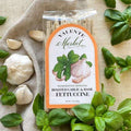 Roasted Garlic & Basil Fettuccine Sold by The Olive Tap: A package of fettucine, surrounded by fresh garlic and basil on a beige canvas material.