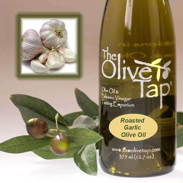 A bottle of Roasted Garlic Olive Oil from The Olive Tap next to olives and leaves with an inset showing bulbs of fresh garlic.