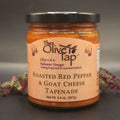 A jar of Roasted Red Pepper and Goat Cheese Tapenade from The Olive Tap with red and green decorative crinkle paper behind the jar, against a gray background and surface.
