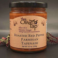 A jar of Roasted Red Pepper Parmesan Tapenade from The Olive Tap with red and green decorative crinkle paper behind the jar against a gray background and surface.