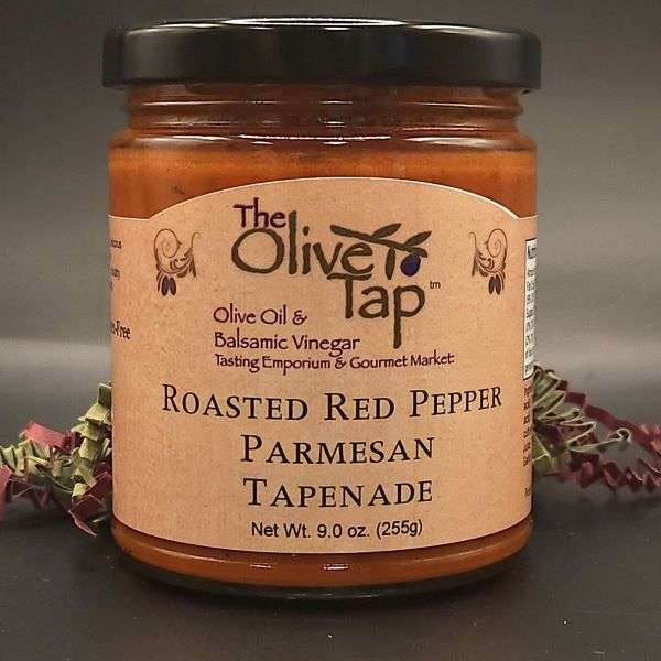 A jar of Roasted Red Pepper Parmesan Tapenade from The Olive Tap with red and green decorative crinkle paper behind the jar against a gray background and surface.