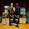 Salad Lovers Gift Box from The Olive Tap: Two large bottles -one olive oil and one vinegar - two smaller bottles of olive oil and two of vinegar, a jar of herbal salt, peppercorns in a grinder, and two metal pouring spouts in front of olive branches and purple grapes on a wooden surface.