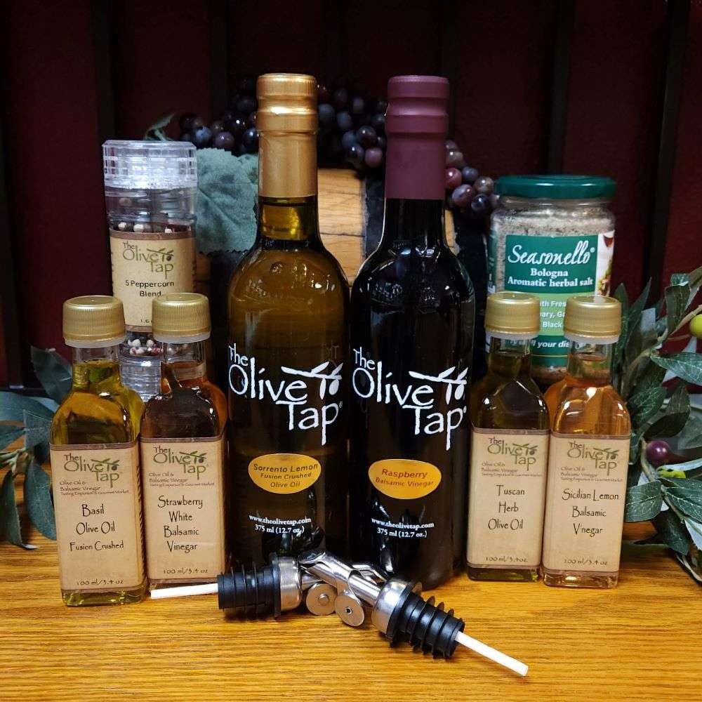 Salad Lovers Gift Box from The Olive Tap: Two large bottles -one olive oil and one vinegar - two smaller bottles of olive oil and two of vinegar, a jar of herbal salt, peppercorns in a grinder, and two metal pouring spouts in front of olive branches and purple grapes on a wooden surface.
