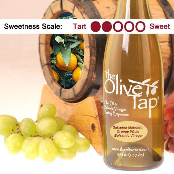 A bottle of Satsuma Mandarin Orange White Balsamic Vinegar from The Olive Tap surrounded by grapes in front of a wooden barrel with mandarins inside, and a sweetness scale indicating a two on a light background.