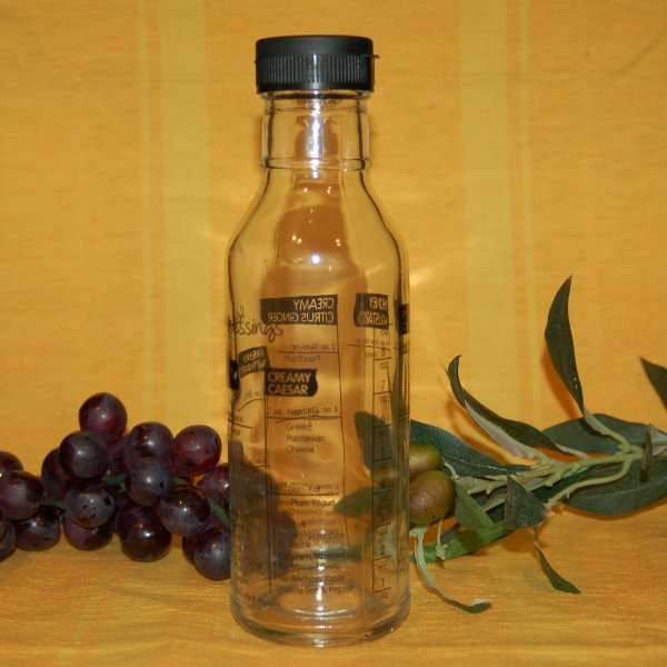 Salad Dressing Shaker & Maker from The Olive Tap: A clear glass bottle with a black cap and writing on it next to grapes, olives, and leaves against a yellow background.