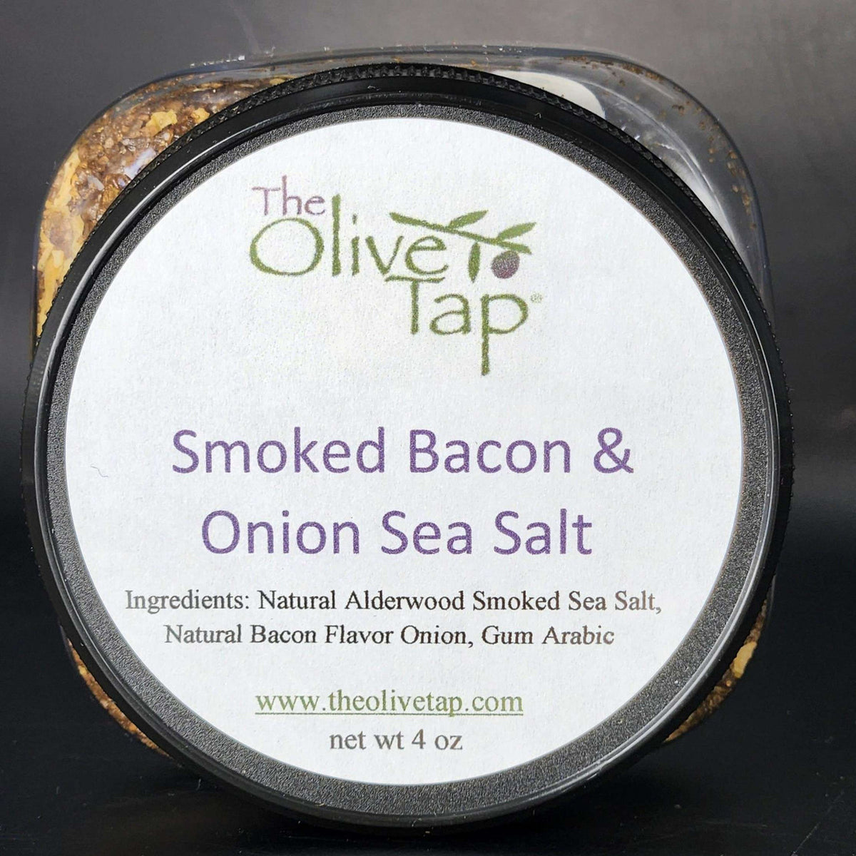Smoked Bacon and Onion Sea Salt In Jar from The Olive Tap in a jar with a white label that includes The Olive Tap logo, product name, and ingredients list, with the jar in front of a gray background.