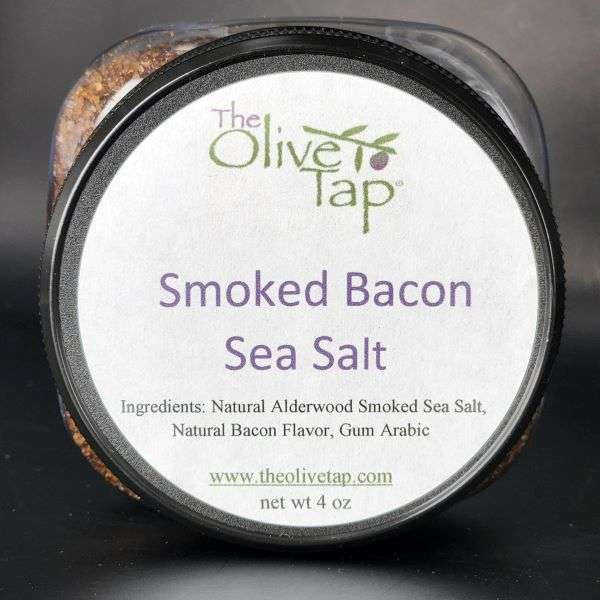Smoked Bacon Sea Salt in a jar from The Olive Tap with a white label that includes The Olive Tap logo with an olive branch, product name, and ingredients list, with the jar in front of a gray background.