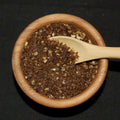 Smoked Bacon and Onion Sea Salt from The Olive Tap: Brown pebble-sized chunks of sea salt with fewer white chunks in a wooden bowl with a wooden spoon inside of it against a black background.