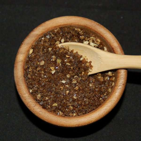 Smoked Bacon and Onion Sea Salt from The Olive Tap: Brown pebble-sized chunks of sea salt with fewer white chunks in a wooden bowl with a wooden spoon inside of it against a black background.