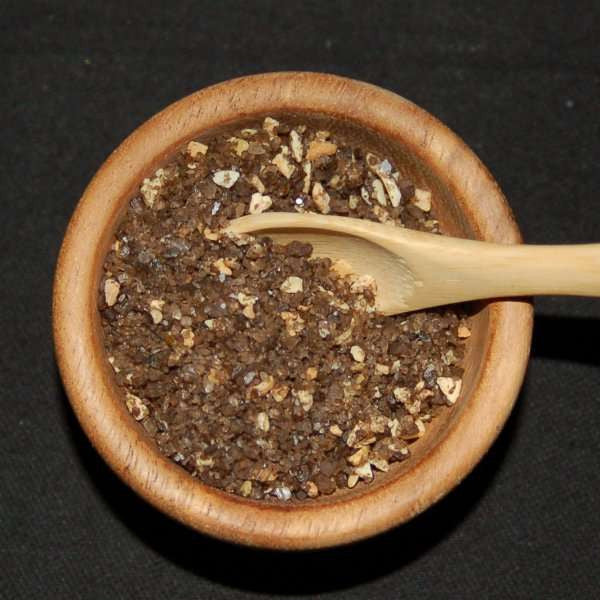Smoked Garlic Sea Salt from The Olive Tap: Pebble-sized chunks of brown sea salt with white chunks in a wooden bowl with a wooden spoon inside of it on a black background.