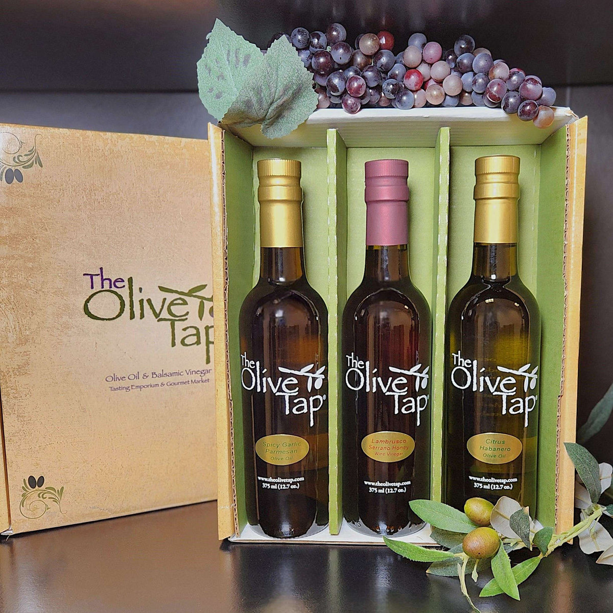 Some Like It Hot Trio Gift Box from The Olive Tap: A decorative Olive Tap box with three bottles inside of olive oils and vinegar, surrounded by decorative grapes and olives.