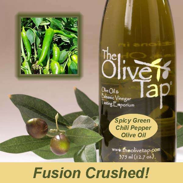A bottle of Spicy Green Chili Pepper Olive Oil from The Olive Tap next to olives and leaves with an inset showing green chilis on a light background with the words "fusion crushed!" at the bottom.