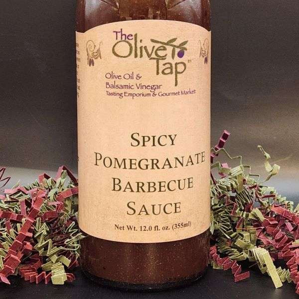 A bottle of Spicy Pomegranate Barbecue Sauce from The Olive Tap surrounded by red and green crinkle paper against a gray background.
