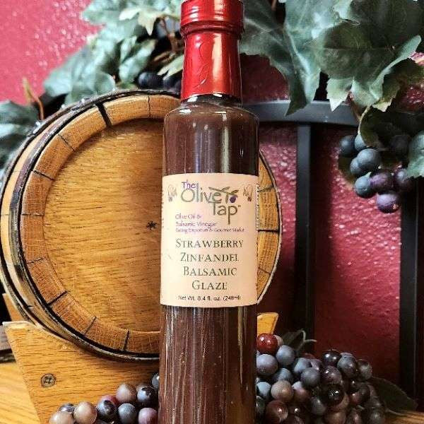 A bottle of Strawberry Zinfandel Balsamic Glaze from The Olive Tap in front of a decorative wooden barrel, purple grapes and their leaves, and a red wall in the background.