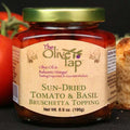 A jar of Sun-Dried Tomato and Basil Bruschetta Topping from The Olive Tap with a fresh tomato to the left and a piece of fresh bread to the right, all on a black surface.