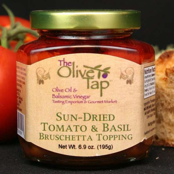 A jar of Sun-Dried Tomato and Basil Bruschetta Topping from The Olive Tap with a fresh tomato to the left and a piece of fresh bread to the right, all on a black surface.
