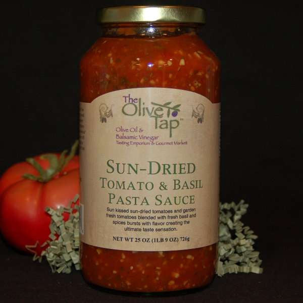 A jar of Sun-Dried Tomato and Basil Pasta Sauce from The Olive Tap with a fresh tomato on the left side and green decorative crinkle paper behind the jar against a black background.