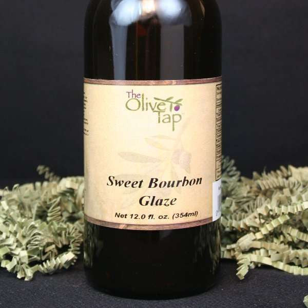 A bottle of Sweet Bourbon Glaze from The Olive Tap, surrounded by green decorative crinkle paper against a black background and surface.