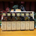 Tap's Sweet Sensations Sampler with Standard Trim from The Olive Tap: Bottles of olive oil and vinegar lined up in a wooden box with a bow on a wooden surface in front of a decorative wooden barrel with olives, leaves, and grapes.