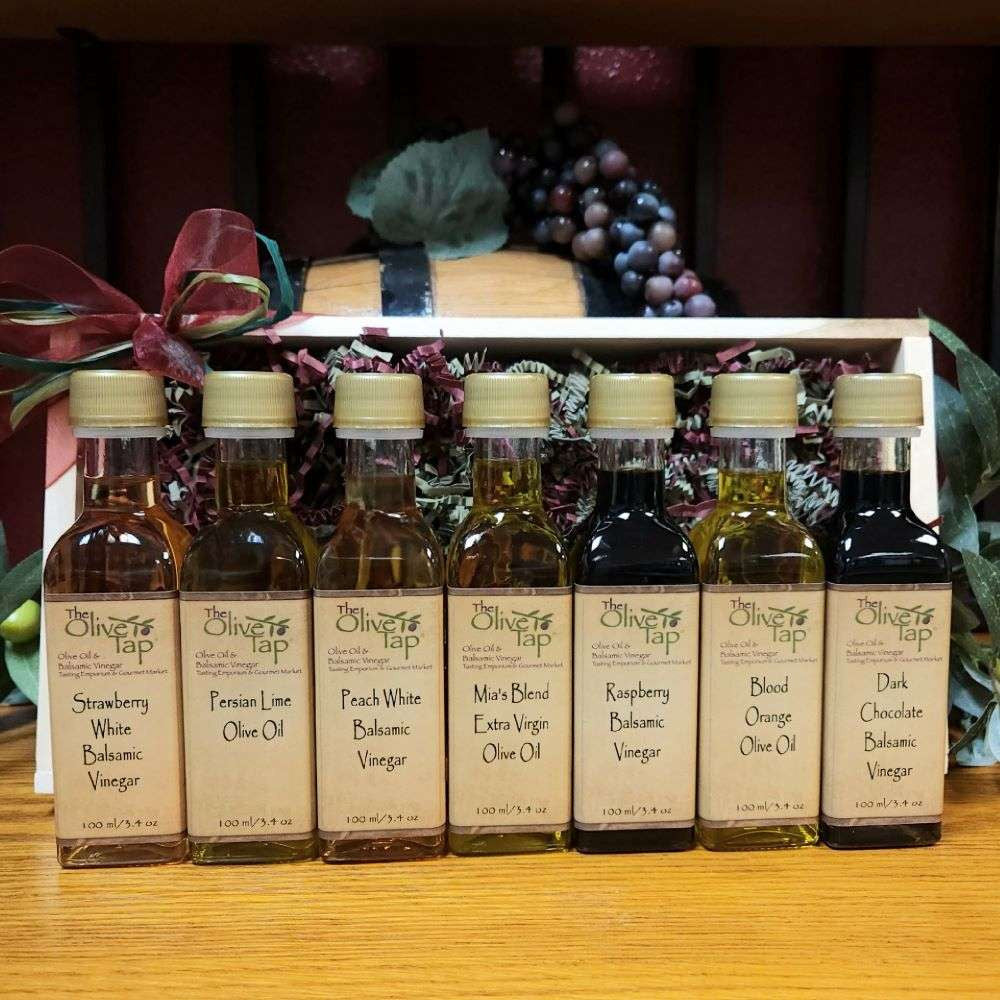 Tap's Sweet Sensations Sampler with Standard Trim from The Olive Tap: Bottles of olive oil and vinegar lined up in a wooden box with a bow on a wooden surface in front of a decorative wooden barrel with olives, leaves, and grapes.