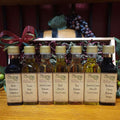 Sweet and Savory Sampler with Standard Trim from The Olive Tap: Bottles of olive oil and vinegar in front of a wooden box with a bow and a wooden barrel with grapes and olives and leaves in the background on a wooden surface.