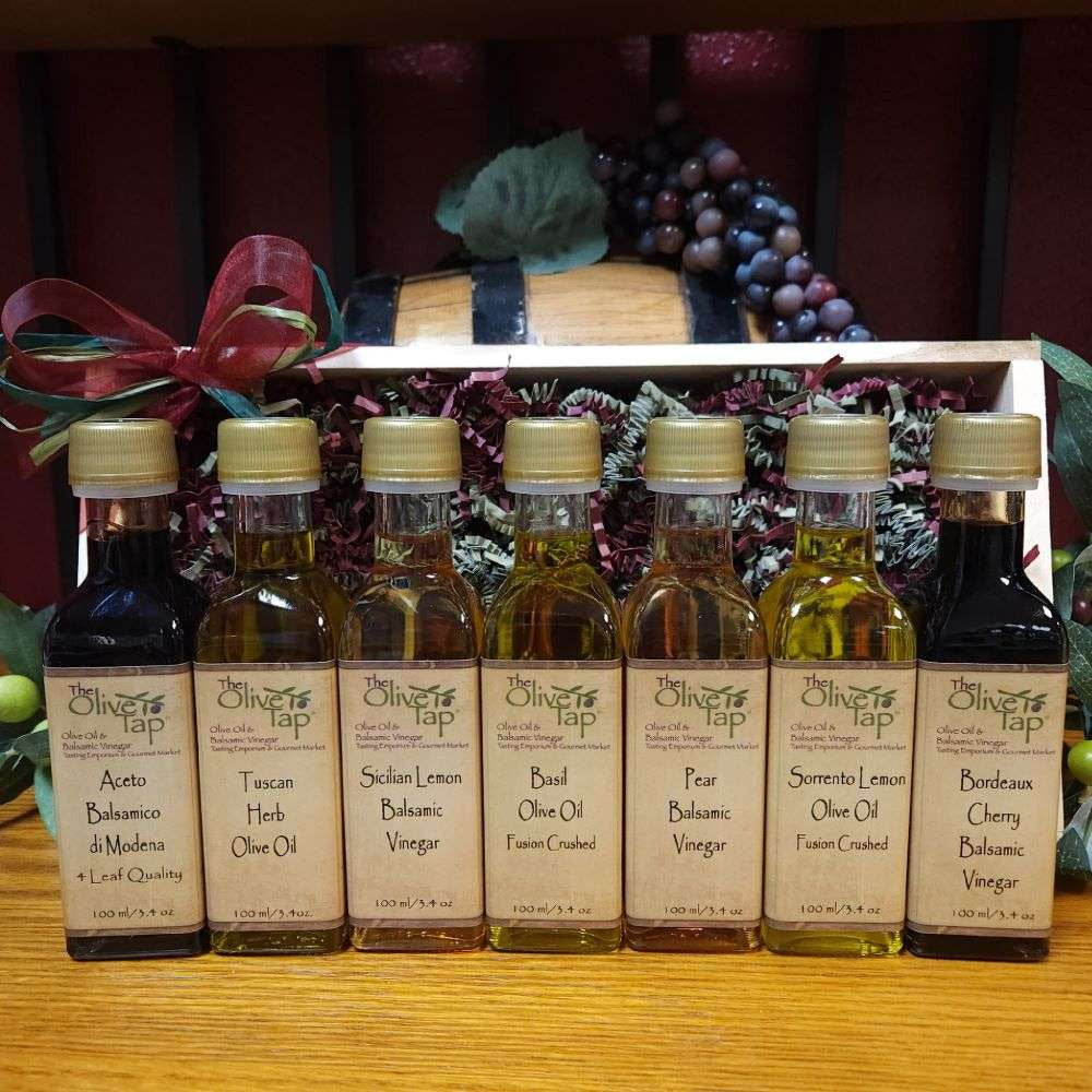 Sweet and Savory Sampler with Standard Trim from The Olive Tap: Bottles of olive oil and vinegar in front of a wooden box with a bow and a wooden barrel with grapes and olives and leaves in the background on a wooden surface.