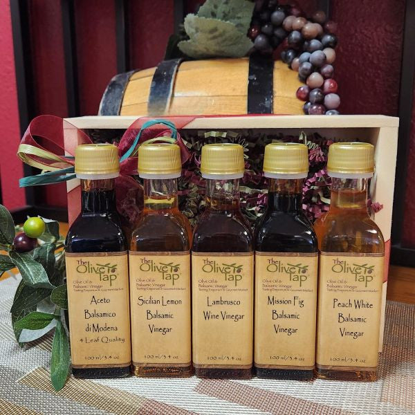 Tap's Tops Vinegar Sampler from The Olive Tap: Five bottles of vinegar lined up in a wooden box with a bow in front of a decorative wooden barrel with olives, leaves, and grapes.