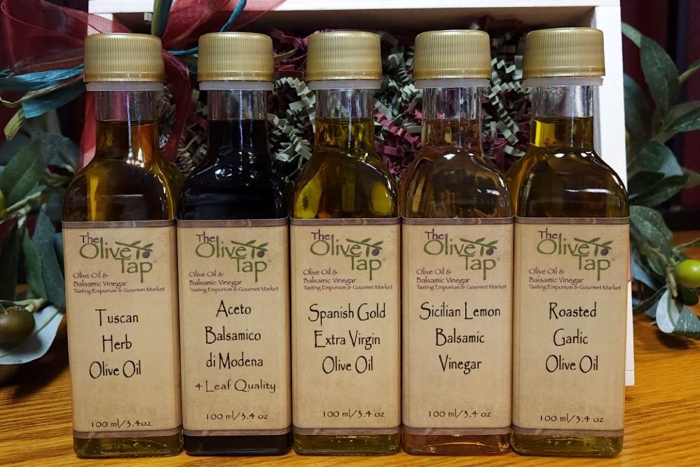 Five bottles of olive oil and vinegar from The Olive Tap on a wooden surface with a decorative background including olives and leaves.