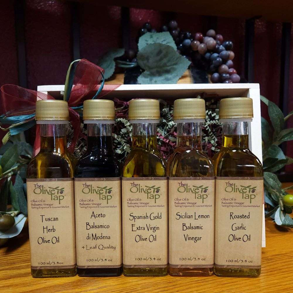 Tap's Tops Sampler from The Olive Tap: Bottles of olive oil and vinegar lined up in a wooden box with a bow on a wooden surface in front of a decorative wooden barrel with olives, leaves, and grapes.