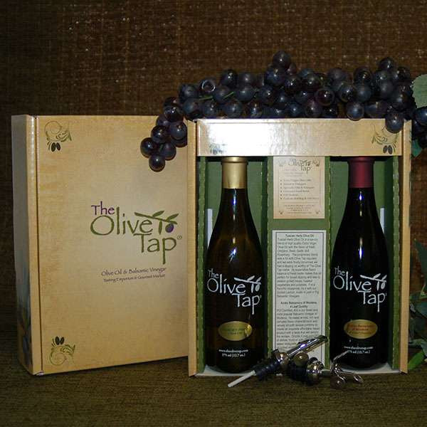 Taste of Italy Gift Box from The Olive Tap: one bottle of olive oil and one bottle of balsamic vinegar in a decorative Olive Tap box with purple grapes on top of it, divided by a piece of cardboard with a description of each product, and two metal pour spouts crossed in front of the box.