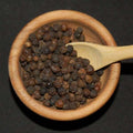 Tellicherry Peppercorns from The Olive Tap in a wooden bowl with a wooden spoon in it against a black background.
