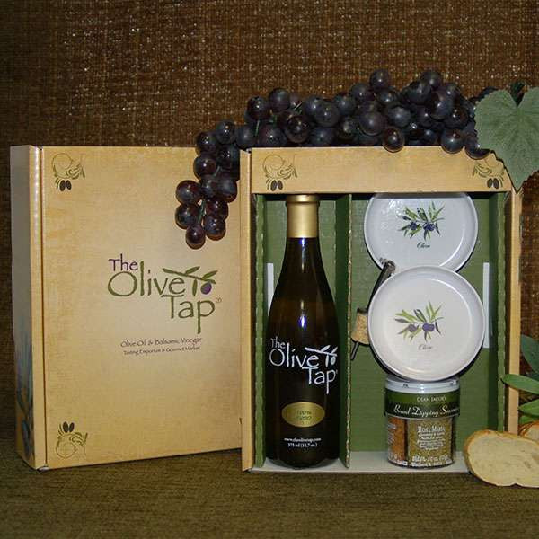 The Bread Dipper Gift Box from The Olive Tap: An Olive Tap gift box with a bottle of olive oil, bread dipping plates, jar of seasoning, and olive oil pour spout inside with grapes on top of the box and a slice of bread next to the box.