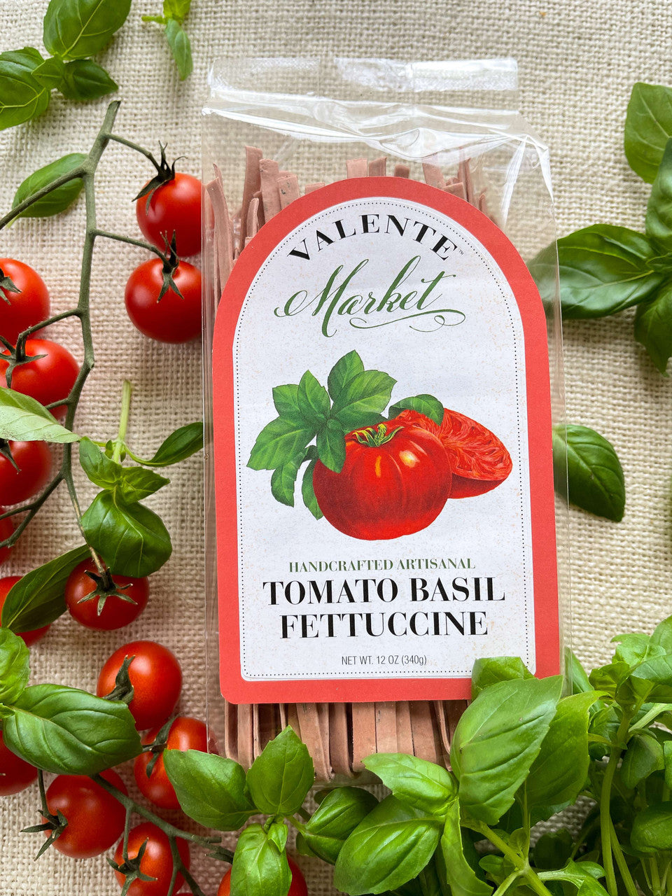 A package of Tomato Basil Fettuccine from The Olive Tap on a light canvas background with small tomatoes on a vine and fresh basil.