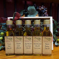 Tap's Tops Oil Sampler from The Olive Tap: Five bottles of olive oil lined up in a wooden box with a bow on a wooden surface in front of a decorative wooden barrel with olives, leaves, and grapes.