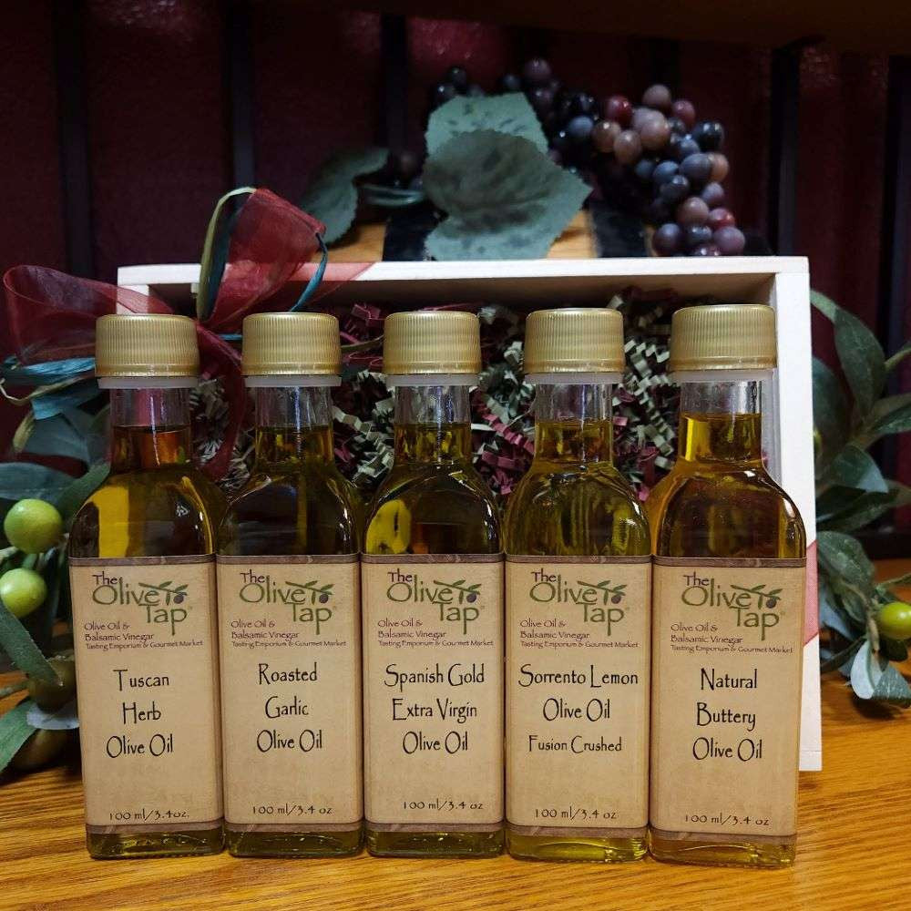 Tap's Tops Oil Sampler from The Olive Tap: Five bottles of olive oil lined up in a wooden box with a bow on a wooden surface in front of a decorative wooden barrel with olives, leaves, and grapes.