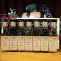 Our Tour of Italy Gift Box with Standard Trim from The Olive Tap: Bottles of olive oil and vinegar lined up in a wooden box with a bow on a wooden surface in front of a decorative wooden barrel with olives, leaves, and grapes.