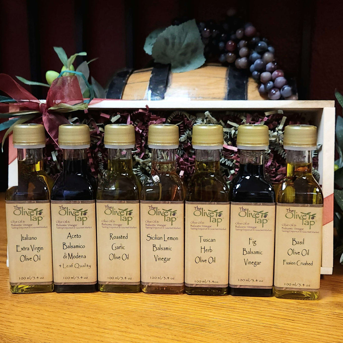 Our Tour of Italy Gift Box with Standard Trim from The Olive Tap: Bottles of olive oil and vinegar lined up in a wooden box with a bow on a wooden surface in front of a decorative wooden barrel with olives, leaves, and grapes.