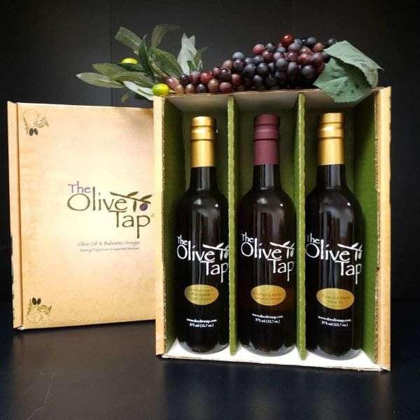 The Olive Tap Trio Gift Box from The Olive Tap: An Olive Tap gift box with three bottles inside and grapes on top of the box on a dark background and surface.