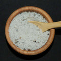 Truffle Sea Salt by The Olive Tap: A wooden bowl and wooden spoon full of white sea salt with dark flakes mixed in against a black background.