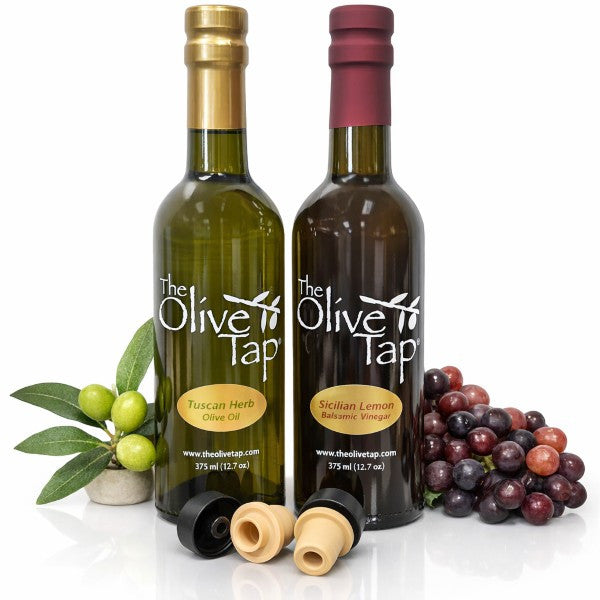 A bottle of The Olive Tap Tuscan Herb Olive Oil and of Sicilian Lemon Balsamic Vinegar  with two pour spouts  and decorateive olives and grapes on a white background