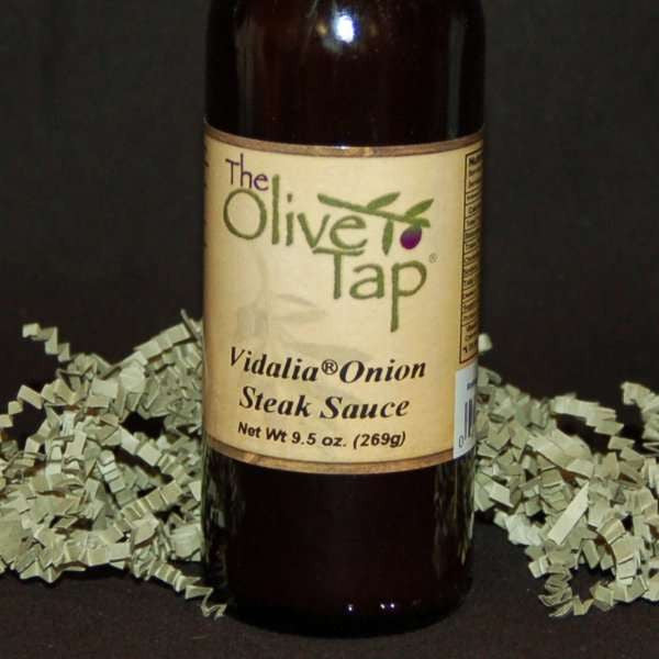 A bottle of Vidalia Onion Steak Sauce from The Olive Tap, surrounded by decorative green crinkle paper against a black background.