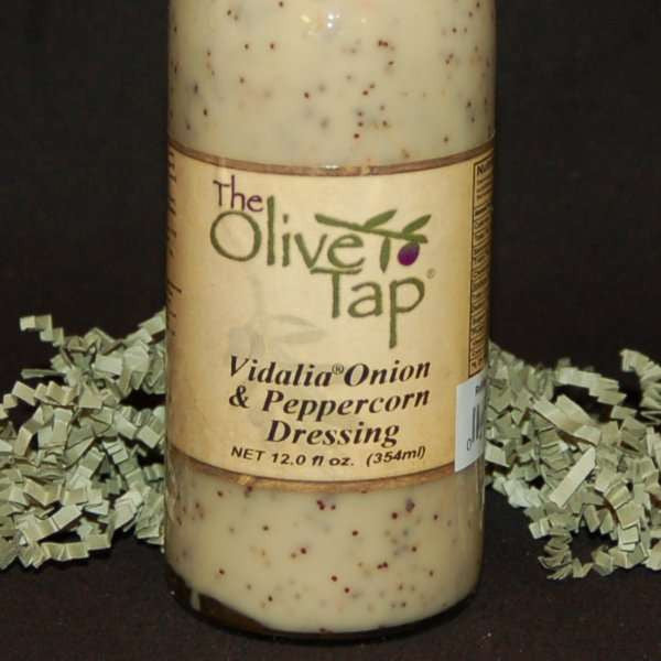 A glass bottle of Vidalia Onion & Peppercorn Dressing from The Olive Tap, surrounded by green decorative crinkle paper against a black background.