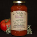 A jar of Vodka Pink Peppercorn Pasta Sauce from The Olive Tap with a fresh tomato on the left side and green decorative crinkle paper behind the jar against a black background.
