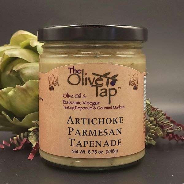 A jar of Artichoke Parmesan Tapenade from The Olive Tap with an artichoke and red and green decorative crinkle paper in the background, against a gray background and on a black surface.