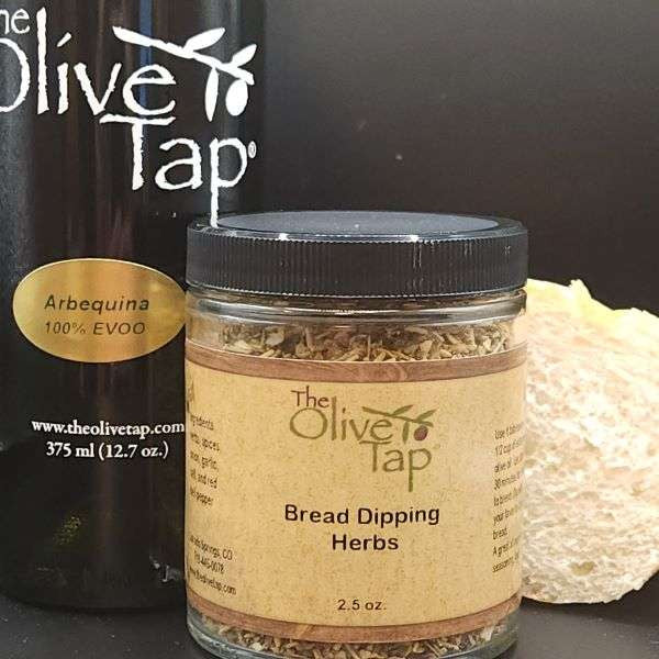 A jar of Bread Dipping Herbs from The Olive Tap next to a bottle of olive oil and bread on a dark background.