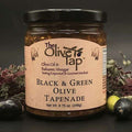A jar of Black and Green Olive Tapenade from The Olive Tap surrounded by black and green olives, red and green decorative crinkle paper, against a gray background and on a black surface.