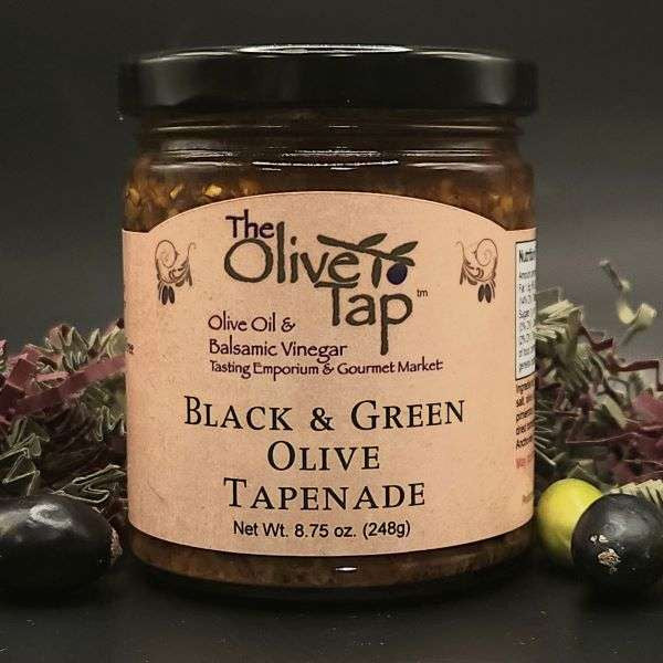 A jar of Black and Green Olive Tapenade from The Olive Tap surrounded by black and green olives, red and green decorative crinkle paper, against a gray background and on a black surface.