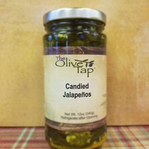 A jar of Candied Jalapeños from The Olive Tap on a plaid surface against a yellow wall.
